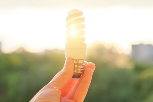 Energy saving fluorescent light bulb, hand holding lamp, evening sunset sky background.