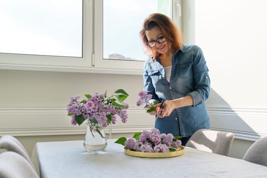 Mature woman making bouquet of lilac branches at home at table in living room