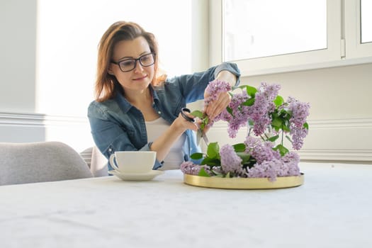 Mature woman making bouquet of lilac branches at home at table in living room