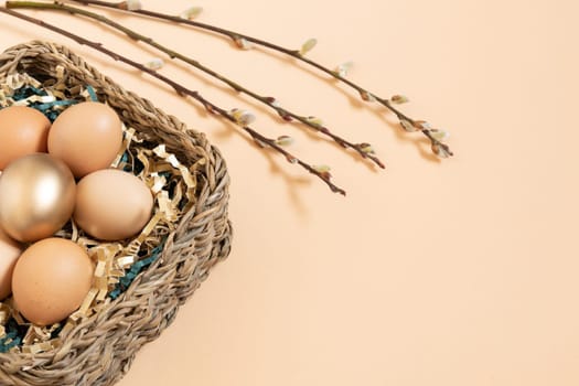 Easter eggs natural and gold in basket with gold and green paper filler . Branch of willow catkins. Light pastel orange background with copy space.