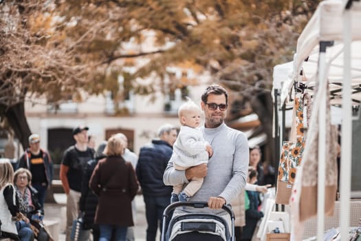 Father walking carrying his infant baby boy child and pushing stroller in crowd of people visiting Sunday flea market in Malaga, Spain