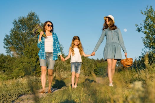 Happy mother and two daughters holding hands walking along rural country road with wildflowers, basket of berries. Sunny summer day, sunset.