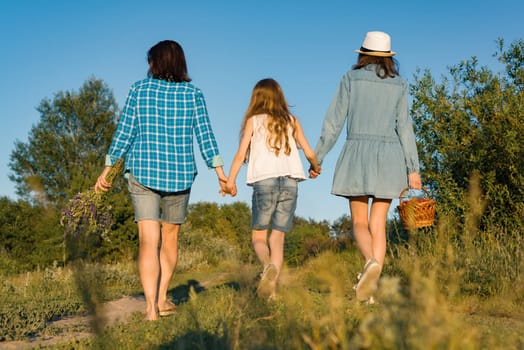 Happy mother and two daughters holding hands walking along rural country road with wildflowers, basket of berries. Sunny summer day, sunset, back view.