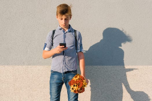 Outdoor portrait of teenage boy with bouquet of flowers, reading text on smartphone, gray wall background copy space.