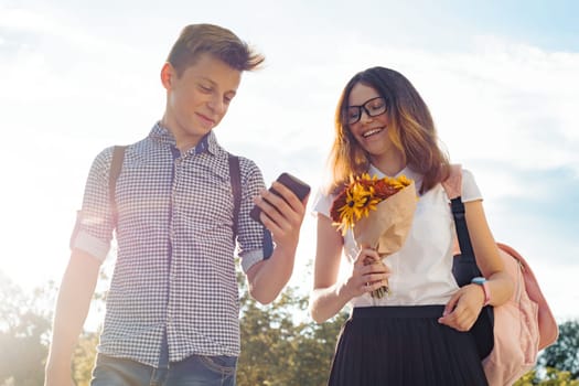 Boy and girl teenagers walking outdoor, couple smiling and talking. Sunny summer day, girl with bouquet of flowers.