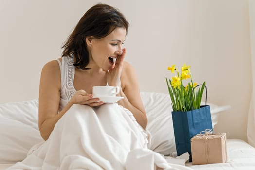 Middle aged woman is pleased with gift, bouquet of flowers, sitting in morning in bed with cup of coffee. Emotion of happiness, joy, surprise.