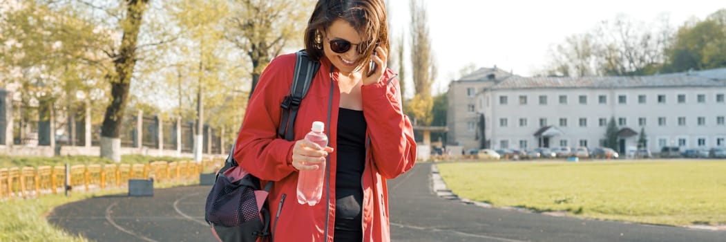 Portrait sporty mature woman at the stadium, panoramic banner, in sports clothes for training, with bottle of water, talking on the phone.