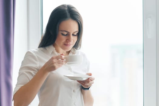 Portrait of young businesswoman in white shirt with cup of coffee, smiling woman enjoying her morning aromatic coffee, office in skyscraper cloudy day, copy space.