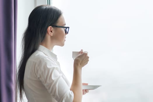 Portrait of young businesswoman in white shirt glasses with cup of coffee, woman looking out the window, office in skyscraper, cloudy day.