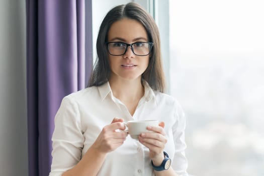 Portrait of young businesswoman in white shirt wearing glasses with cup of coffee, posing smiling woman looking into the camera, office in skyscraper, cloudy day.