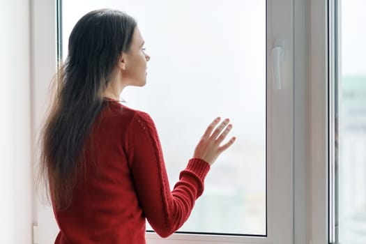 Young woman in red sweater looking out the window, female put her hands on the window glass, side view, window in skyscraper in morning on cloudy day.