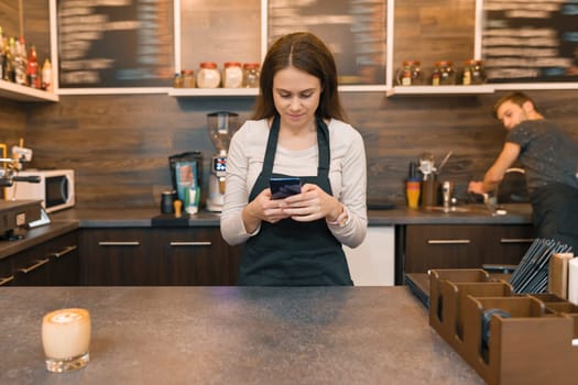Young woman in apron coffee shop worker at bar counter, taking order on smartphone.