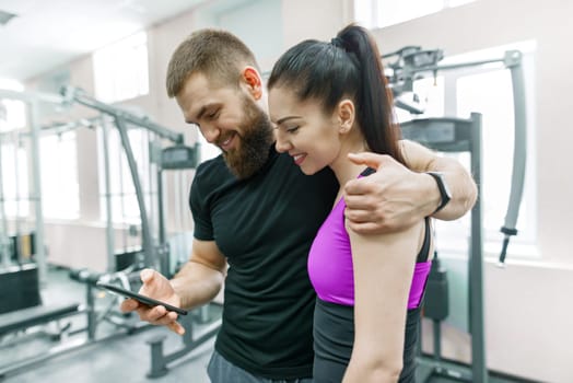 Young woman talking with personal trainer in gym, looking in smartphone and discussing. Fitness, sport, training, people, healthy lifestyle concept.
