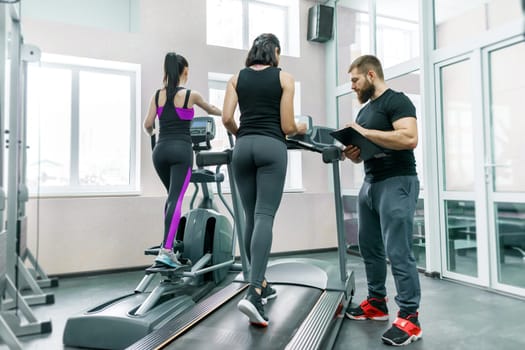 Young smiling fitness women with personal trainer an adult athletic man on treadmill in the gym. Sport, teamwork, training, healthy lifestyle concept.