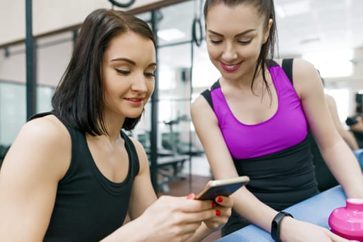 Two young sports women in gym talking smiling with fitness mats and looking at mobile phone. Training, teamwork, healthy lifestyle concept