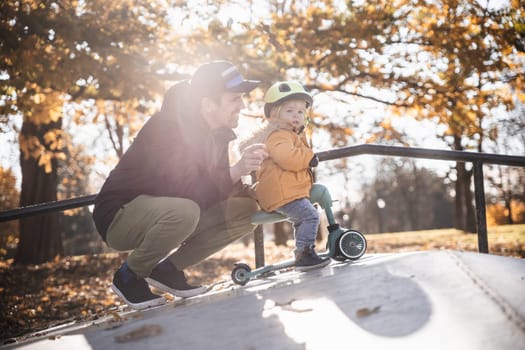 Father supervises his fearless small toddler boy while riding baby scooter outdoors in urban skate park. Child wearing yellow protective helmet.