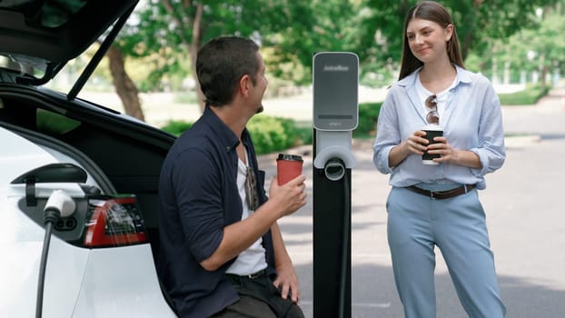 EV car recharging electricity for battery by lovey couple traveling during their road trip travel by eco friendly electric vehicle in national park and greenery forest on holiday. Exalt