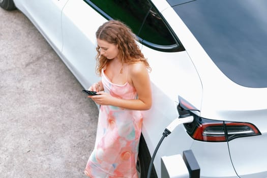 Aerial top view eco-friendly woman recharge electric vehicle from EV charging station, using EV technology utilization for tracking energy usage to optimize battery charging on smartphone. Synchronos