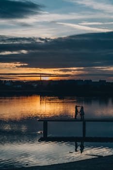 Silhouettes of young couple holding hands standing on Bagry lake pier at sunset, Poland