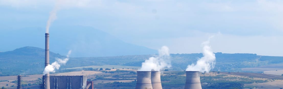 Smoking chimneys of a thermal power plant against the backdrop of fields and mountains, panoramic photo in blue tones