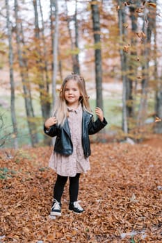 Little smiling girl stands under falling dry leaves in the autumn forest. High quality photo