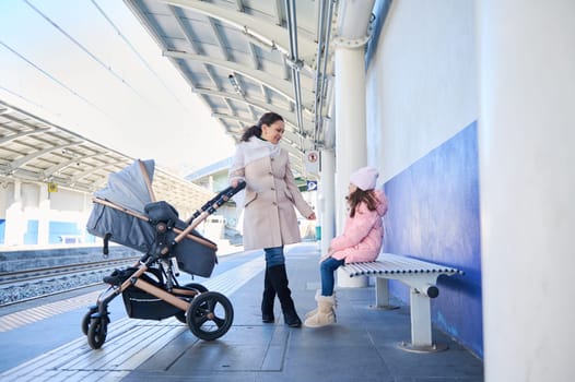 Full length portrait of a mother with children waiting train on the railway station platform. Pretty woman commuter pushing baby pram and talking to her daughter while travelling together by train