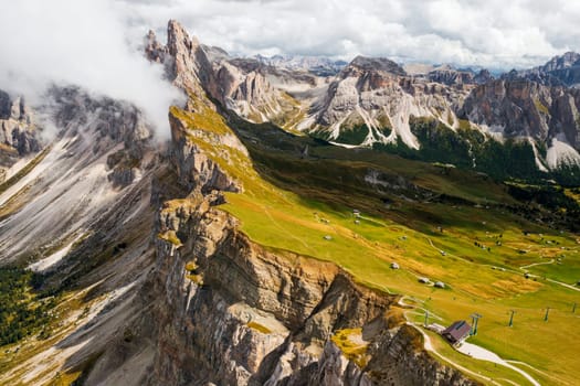 Tourist observation point against Seceda mountain with rocky peaks and green grass under cloudy sky. Scenic Italian Alps in summer aerial view