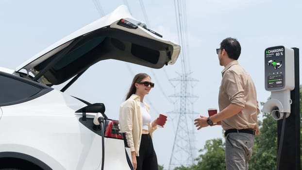 Young couple with coffee recharge EV car battery at charging station connected to electrical industrial power grid tower. Couple with shopping and travel using eco electric car lifestyle. Expedient