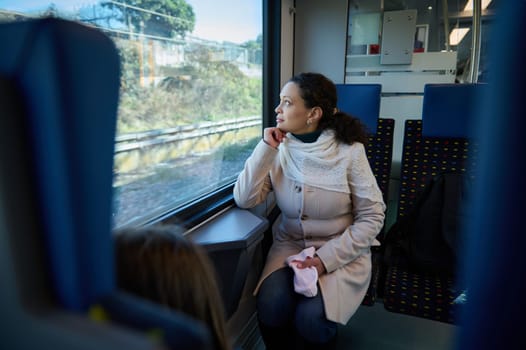 Thoughtful young woman commuter travelling by train, dreamily looking out the window, sitting on passenger seat, enjoying comfortable trip from one city to other. Urban public rail road transportation