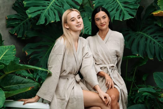 Tropical and exotic spa garden with bathtub in modern hotel or resort with young two women in bathrobe enjoying leisure and wellness lifestyle surrounded by lush greenery foliage background. Blithe