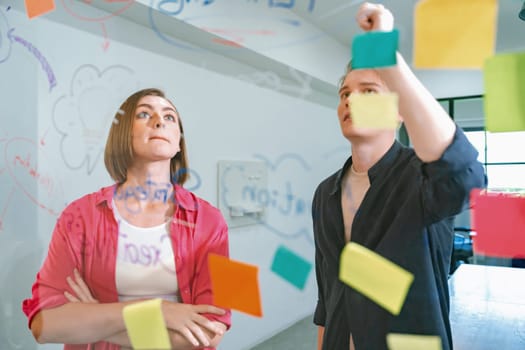 Couple of businesspeople discussing and brainstorming about solving marketing problem in front of glass board at business meeting by using mind map and sticky notes. Working together. Immaculate.