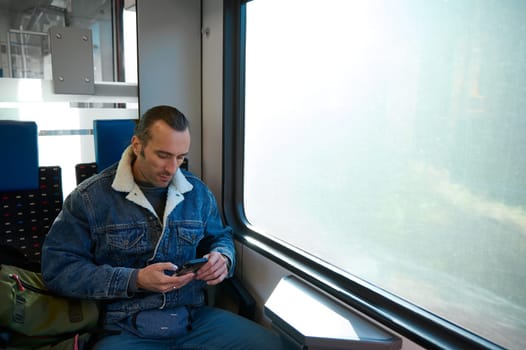 Caucasian handsome male passenger, commuter using smartphone, checking mobile app, planning route while travelling by comfortable city train. Online communication. Internet. Urban transportation