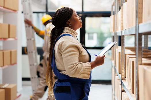 Warehouse supervisor looking at parcels and taking notes in storage room. African american logistics manager holding clipboard with order checklist and planning distribution