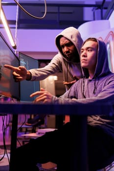 Confused hacker pointing at computer screen and asking criminal about password cracking technique. Diverse young men in hoods planning phishing attack while breaking law together
