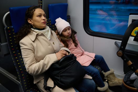 Portrait of diverse people travelling by train. Young woman thoughtfully looking away, sitting near her daughter, commuting from work to home using railroad high speed transport.