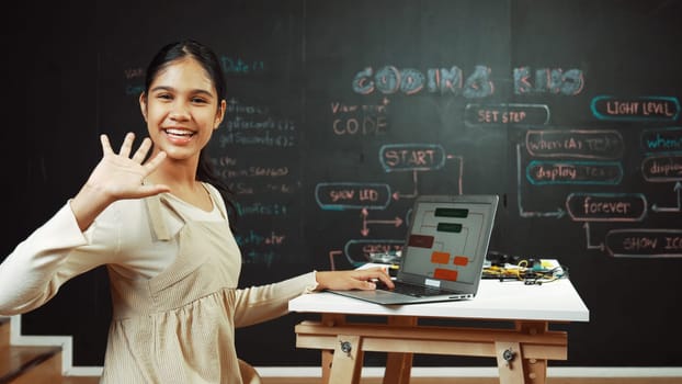 Smart highschool girl waving to camera while programing code at blackboard with engineering code or prompt written. Academic student working on laptop or writing program and greeting. Edification.