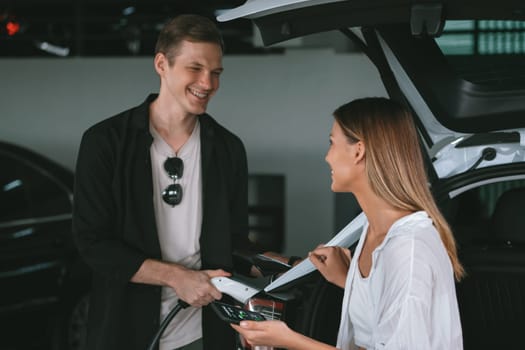 Young couple travel with EV electric car to shopping center parking lot charging in downtown city showing urban sustainability lifestyle by green clean rechargeable energy of electric vehicle innards