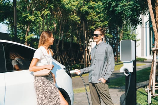 Young couple travel with EV electric car charging in green sustainable city outdoor garden in summer shows urban sustainability lifestyle by green clean rechargeable energy of electric vehicle innards