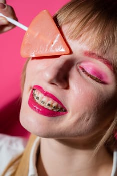 Portrait of a young woman with braces and bright makeup eating a lollipop on a pink background. Vertical photo