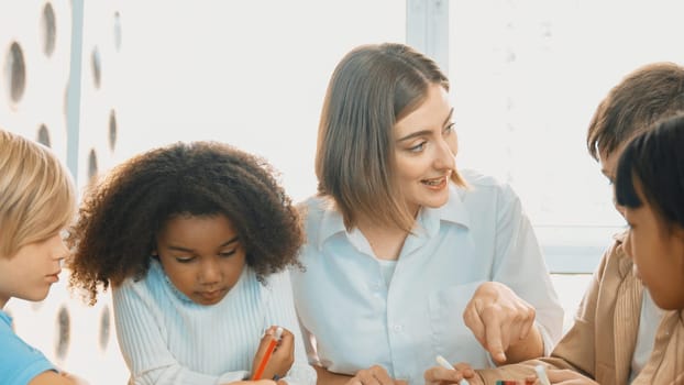 Professional caucasian teacher telling story to diverse student while sitting at table with storybook and colored book. Smart learner listening story while colored picture from instructor. Erudition.