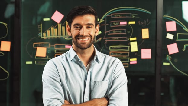 Professional businessman looking at camera while standing with folded arms on glass wall with statistic and sticky notes. Male leader crossing arms while smiling at camera with confident. Tracery