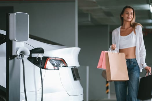 Young woman travel with EV electric car to shopping center parking lot charging in downtown city showing urban sustainability lifestyle by green clean rechargeable energy of electric vehicle innards