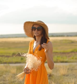 Young woman in an orange dress and a straw hat standing on a field in the rays of the setting sun.