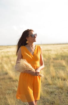 A beautiful brunette girl in an orange dress and sunglasses with a bouquet of field grass enjoys nature in the summer