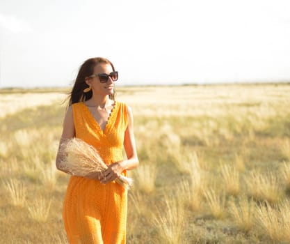 Beauty girl outdoors enjoying nature. A beautiful girl in an orange dress, walks across the field with a bouquet of feather grass, across the steppe, sunlight