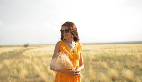 A beautiful brunette girl in an orange dress and sunglasses with a bouquet of field grass enjoys nature in the summer