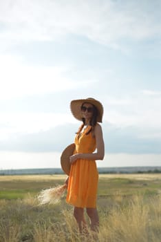 Young woman in an orange dress and a straw hat standing on a field in the rays of the setting sun.