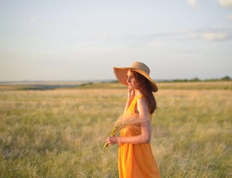 Young woman in an orange dress and a straw hat standing on a field in the rays of the setting sun.