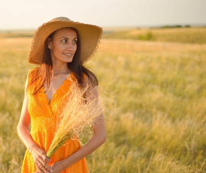 Young woman in an orange dress and a straw hat standing on a field in the rays of the setting sun.