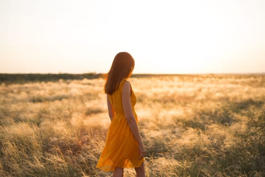 A pretty young girl with long brown hair in an orange dress in a field at sunset. The concept of freedom and nature